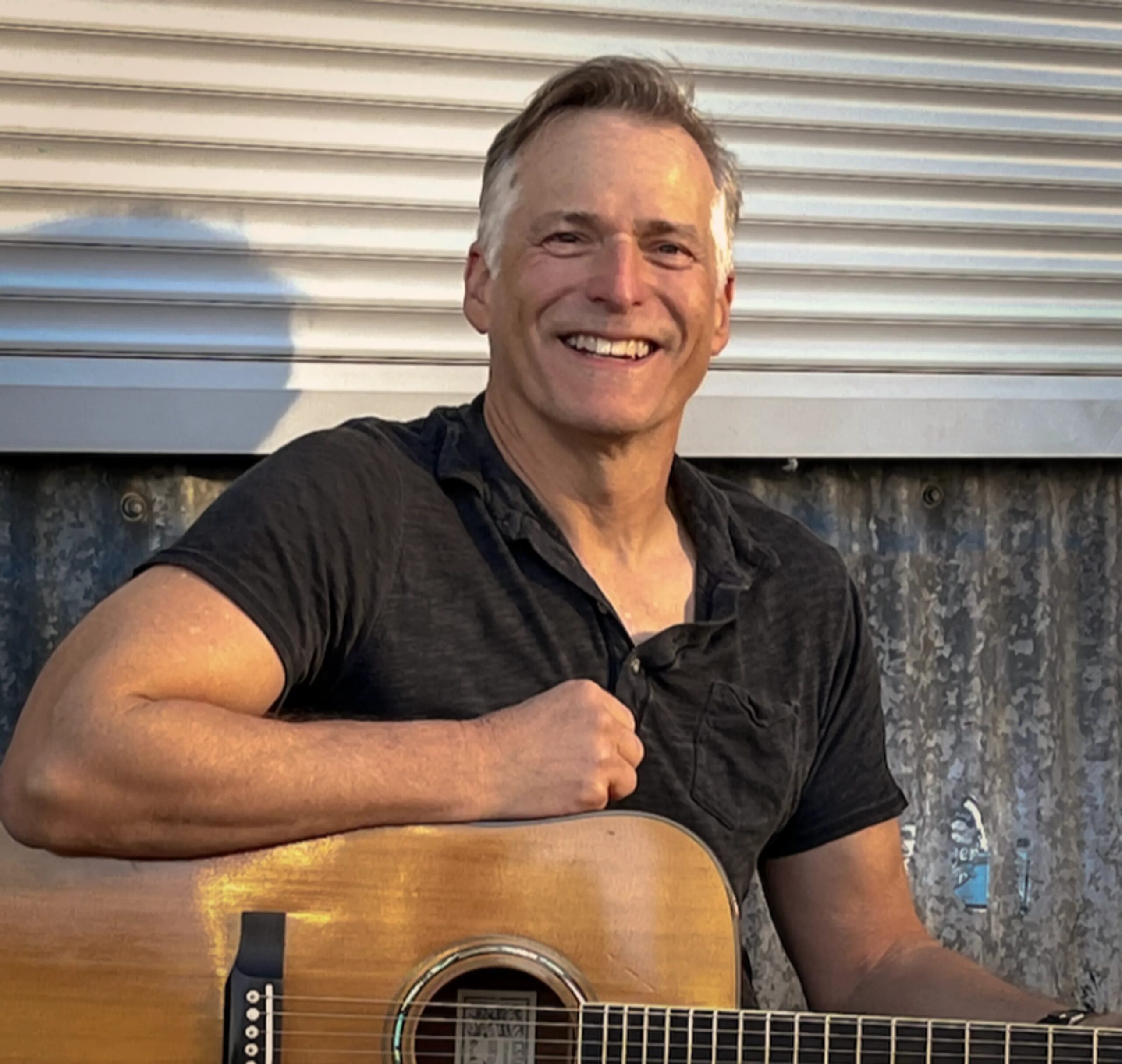 Jeff Standfield smiles while sitting and holding a guitar.